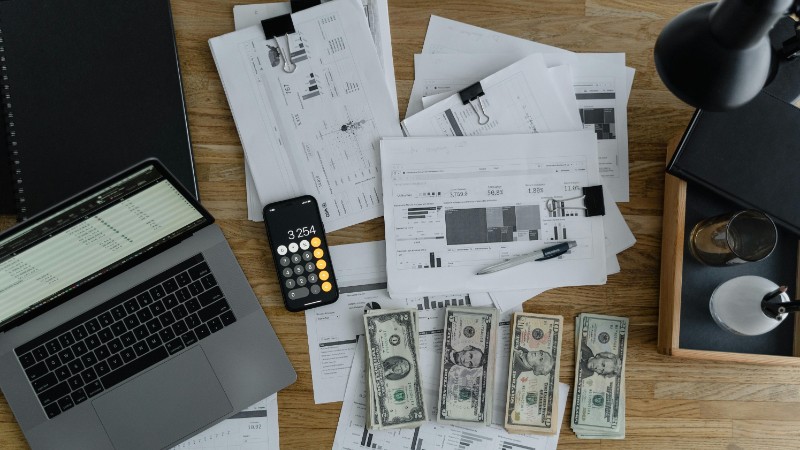 Overhead view of financial documents, cash, and technology on a wooden desk. - pexel