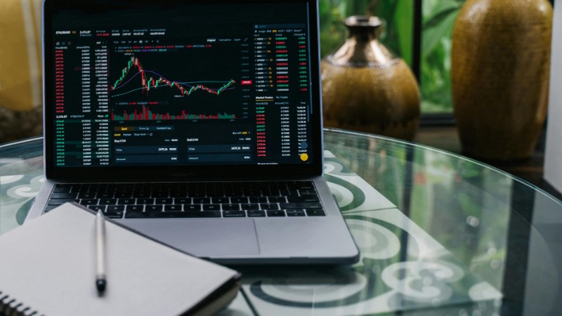 Laptop displaying cryptocurrency stocks and graphs on a glass table with a notepad., tags: stock market - pexel