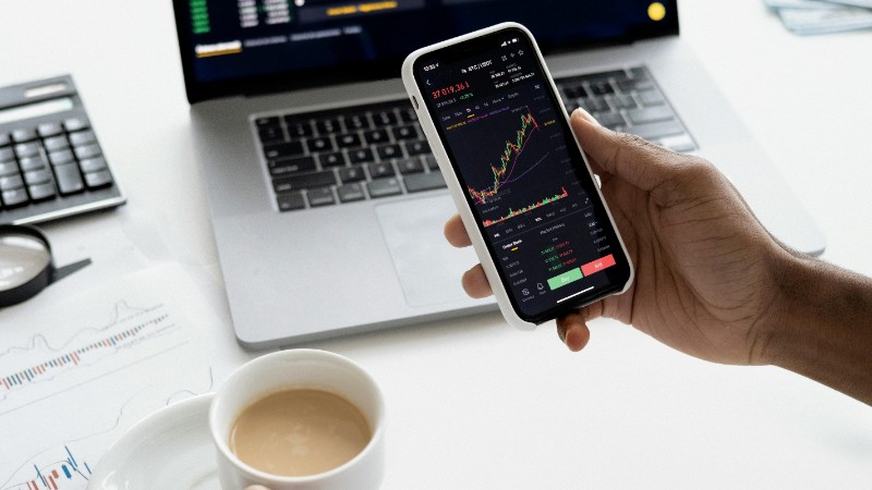 A person trades stocks on a smartphone with a laptop, coffee cup, calculator, and documents in view., tags: wall street 2026 - pexel