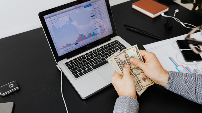 Person counting dollar bills with financial graphs on a laptop screen, symbolizing financial analysis and planning., tags: avoiding common investment - pexel