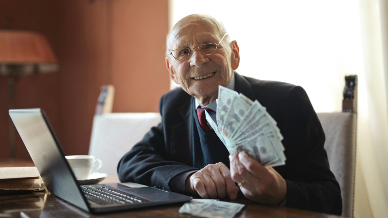 Happy senior businessman showing cash at his desk with a laptop, indoors., tags: economic - pexel