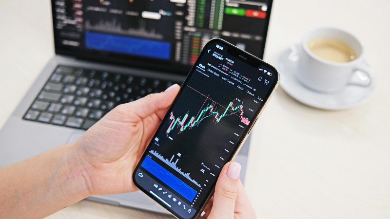 Close-up of hands holding smartphone displaying stock charts with laptop in the background on a desk., tags: federal reserve liquidity - pexel