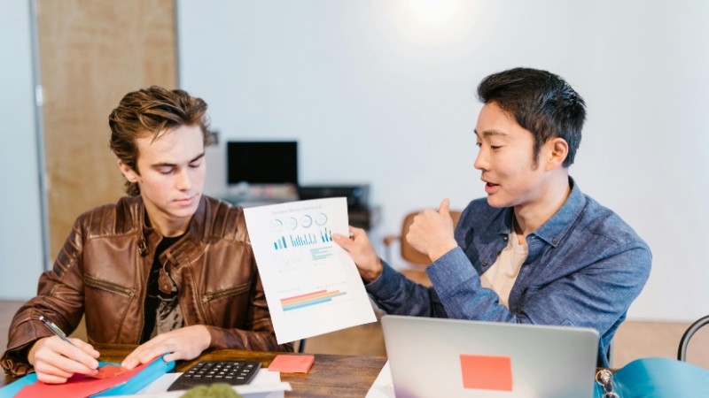 Two men discuss graphs and data at a shared workspace in a modern office setting., tags: retirement young - pexel