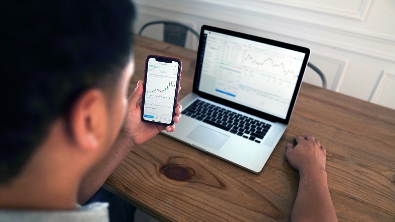 Individual reviewing stock market trends using a laptop and smartphone at a wooden table., tags: investors inflation - pexel