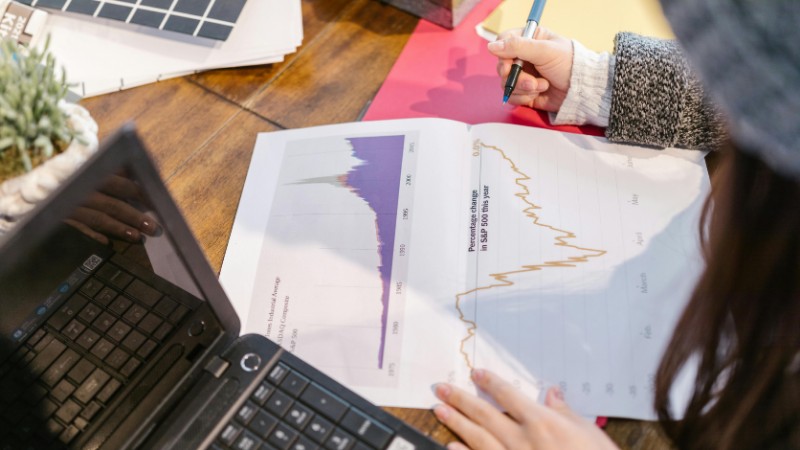 Person reviewing financial graphs and charts while taking notes beside a laptop., tags: capital war s&p 500 - pexel